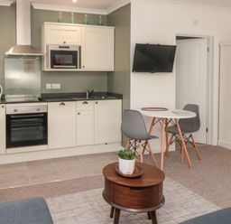A kitchen area with white cabinets and appliances next to a small dining table with two chairs and a wall-mounted TV at Flodden in Crookham near Cornhill-On-Tweed