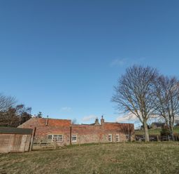 A brick and stone building with multiple windows near a wooden shed and leafless trees on grassy land at Bakers Cottage in Crookham near Cornhill-On-Tweed