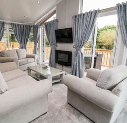 A living room with beige sofas a glass coffee table wall mounted tv and windows with blue curtains at Woodpecker Lodge in Highampton near Sheepwash