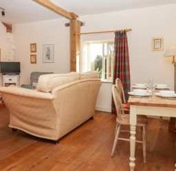 A living room with a dining table set with dishes and glasses a beige sofa wooden chairs a window with plaid curtains and a small television at The Old Dairy in Colyton