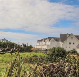 A row of white houses with pitched roofs behind green shrubs and grass at 9 Lon Y Dryw in Trearddur Bay