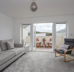 A sitting room with grey sofa and armchair opening onto a small balcony with wooden table and chairs at 9 Lon Y Dryw in Trearddur Bay