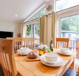 A dining area with a wooden table set with white dishes and bowls glasses bread and fruit next to large windows with curtains at Garuda Calgarth 19 White Cross Bay near Windermere