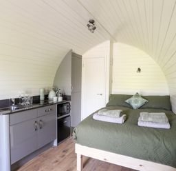 A small room with a bed with green bedding and folded towels a kitchenette with a sink glasses a kettle and microwave at Tyddyn Parthle Pod 1 in Bontnewydd Gwynedd