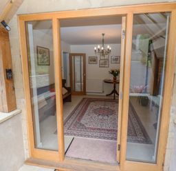 An entrance with glass wooden doors opening into a room with a patterned rug a chandelier wall pictures a wooden bench and a round table with flowers at The Gables in Sheepscombe