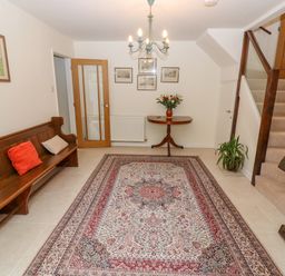A hallway with a wooden bench and cushions a patterned rug a small table with flowers a staircase and framed pictures at The Gables in Sheepscombe