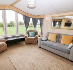 A living room with a grey sofa and two armchairs with cushions a large mirror on the wall and large windows with curtains at Plas Tirion Caravan Llanfairynghornwy near Cemaes Bay