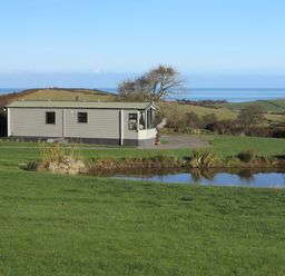 A mobile home near a small pond with green fields and trees in the background at Plas Tirion Caravan Llanfairynghornwy near Cemaes Bay