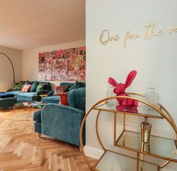 A living room with blue sofas a floor lamp and a wall art with a bar cart and pink rabbit figure at Tipsy Bee in Bowness-On-Windermere