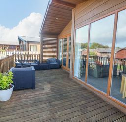 A wooden deck with outdoor seating including chairs and a glass table adjacent to a cabin with large windows at Gressingham 22 in South Lakeland Leisure Village