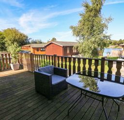 A wooden deck with a chair and glass table overlooking cabins and a lake at Gressingham 22 in South Lakeland Leisure Village