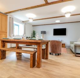 A dining area with a wooden table and benches beside a living room with a sofa and a wall-mounted TV at Budock in Mawnan Smith Near Falmouth