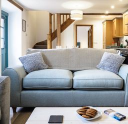 A living room with a blue sofa and pillows with a kitchen and wooden stairs in the background at Kerensa in Mawnan Smith Near Falmouth