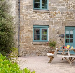 An outdoor patio with a wooden picnic table set for breakfast next to a stone building with blue windows and door at Newlyn in Mawnan Smith Near Falmouth