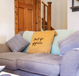 A grey sofa with three cushions and a blanket in a room with a wooden door and staircase at Newlyn in Mawnan Smith Near Falmouth