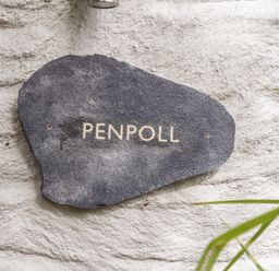 A dark stone plaque with the word Penpoll mounted on a textured white wall with some green leaves in the corner at Penpoll in Falmouth