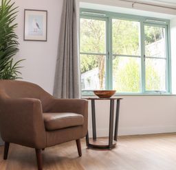A brown armchair next to a small round wooden table with a bowl and a large window with grey curtains and a tall green plant at Penpoll in Falmouth