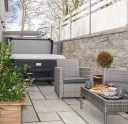 An outdoor patio area with a hot tub a wicker sofa and armchair a glass coffee table with food and drinks and plants in wooden pots at Penpoll in Falmouth
