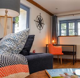 A living room with a blue sofa with patterned pillows a wooden floor lamp a desk with an orange chair and blue framed windows at St Keverne in Mawnan Smith Near Falmouth