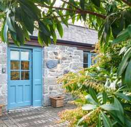 A stone house with a blue door and window framed by green leafy plants at Manacles in Mawnan Smith Near Falmouth