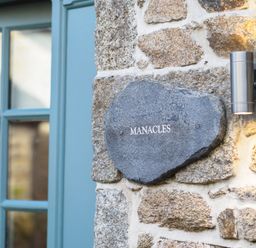 A stone plaque with the word Manacles mounted on a stone wall next to a blue window frame and a wall light at Manacles in Mawnan Smith Near Falmouth