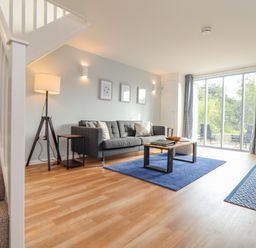 A living room with a gray sofa a wood and metal coffee table a blue rug a floor lamp stairs and sliding glass doors at Helford in Mawnan Smith Near Falmouth