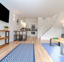 A living room with a wall-mounted television and a wooden shelf a dining table with benches a staircase and a kitchen in the background at Helford in Mawnan Smith Near Falmouth