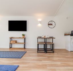 A living room with a wall mounted tv a wooden table a clock two stools a washing machine and coffee maker at Helford in Mawnan Smith Near Falmouth