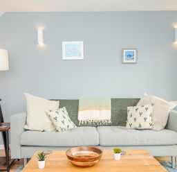 A living room with a grey sofa with cushions and a throw blanket a wooden coffee table with plants a floor lamp and framed pictures on a blue wall at Mousehole in Mawnan Smith Near Falmouth