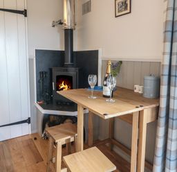 A small wooden dining table with two glasses and a bottle next to a stove with fire in a room with plaid curtains at Coraldie Laggan near Newtonmore