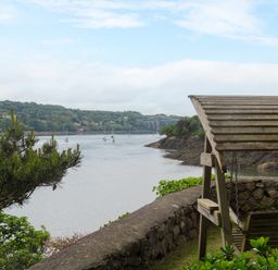 A riverside view with a wooden swing seat under a pergola near a stone wall and boats on the water at Ynys Castell in Menai Bridge