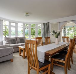 A dining area with wooden table and chairs and a living space with gray sofas in front of large windows at Ynys Castell in Menai Bridge