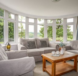 A living room with a sofa, two armchairs, a wooden coffee table, and large windows showing greenery at Ynys Castell in Menai Bridge