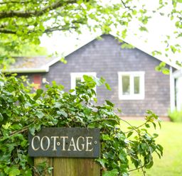 A green leafy bush with a sign that says cottage in front of a dark wooden house with white windows at Henllys Lodge in Newport Pembrokeshire