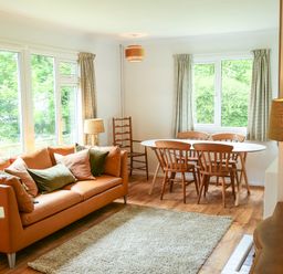 A living room with a brown sofa with cushions a white oval dining table with wooden chairs a floor lamp and windows with curtains at Henllys Lodge in Newport Pembrokeshire