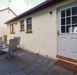 An outdoor patio area with four wicker chairs and a glass table beside a cream house with three small windows and a white door at Lilyvale in Davidstow near Camelford