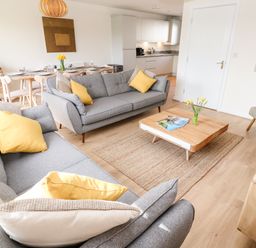 A living room with two grey sofas with yellow and beige cushions a wooden coffee table with magazines and flowers a dining table set with plates and glasses and a wooden sideboard at The Bright House in St Eval near St Columb Major