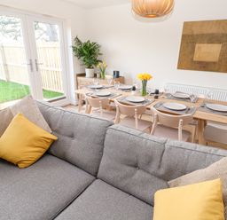 A dining area with a wooden table set for six and a grey couch with yellow and beige cushions at The Bright House in St Eval near St Columb Major