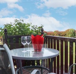 A glass outdoor table with two wine glasses and a red wine bottle holder on a wooden balcony with greenery and blue sky at Lodge Two Country View Park in Graveney