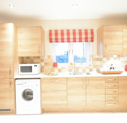 A kitchen with wooden cabinets a washing machine a microwave and a window with a red checkered blind at Red Robin Whepstead in Wickhambrook near Bury St Edmunds