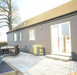 An outdoor patio area with dining table and chairs beside a building with black siding and glass double doors at Red Robin Whepstead in Wickhambrook near Bury St Edmunds