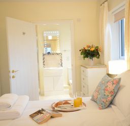 A bedroom with a bed with white bedding and a floral pillow a tray with croissants and orange juice towels a dresser with flowers a lamp and a view into a bathroom at Red Robin, Whepstead in Wickhambrook near Bury St Edmunds
