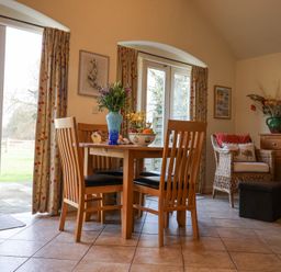 A dining area with a wooden table and chairs a wicker chair a wooden sideboard and flowers in a vase at Romulus in Larling near East Harling