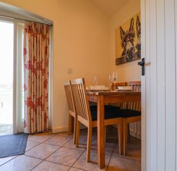 A dining area with a wooden table set with plates and glasses and wooden chairs near a glass door with floral curtains at Remus Larling near East Harling