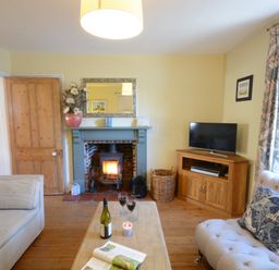 A living room with a fireplace a wooden door a sofa a TV on a wooden cabinet and a coffee table with wine glasses at Pebble Beach Cottage Aldeburgh