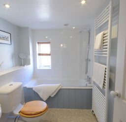 A bathroom with a bathtub a toilet with wooden seat a towel rack and a small window at Pebble Beach Cottage Aldeburgh