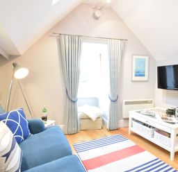 A small living room with a blue sofa pillows and floor lamp near a window with curtains a wall mounted tv and a white coffee table with magazines and baskets at Pied a Mer Aldeburgh