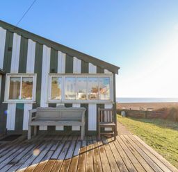 A wooden deck with a bench and chair outside a striped cabin near a grassy area and a beach at Valetta Thorpeness in Thorpeness