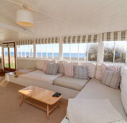 A living room with a sectional sofa striped cushions a wooden coffee table and large windows showing a sea view at Valetta Thorpeness in Thorpeness
