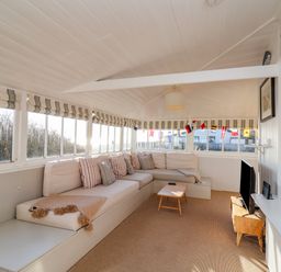A sunroom with a large corner sofa and striped cushions a small wooden table and a TV on a wooden stand at Valetta Thorpeness in Thorpeness